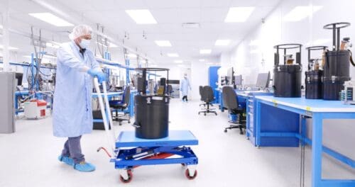 A worker in protective clothing moves a large black container on a blue cart inside a bright, clean laboratory with various equipment and workstations.