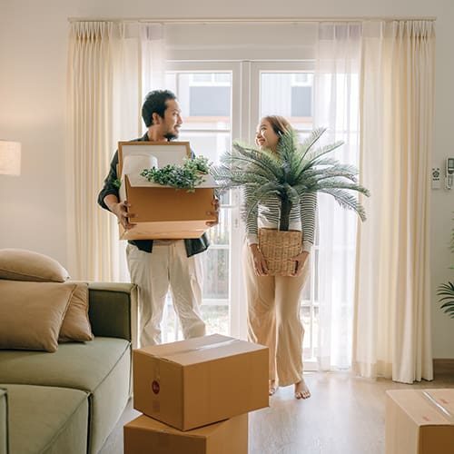 Couple moving into a new home with boxes and plants in a bright living room.